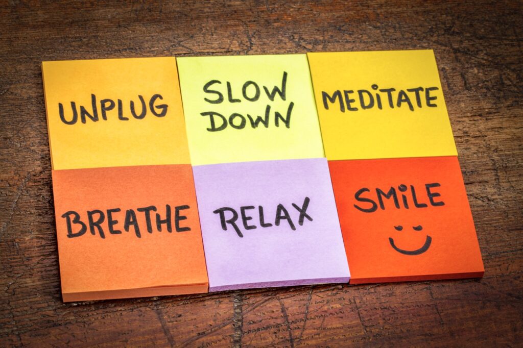Photo of motivational reminders on multi-color sticky notes against grunge wood with the words unplug, slow down, meditate, breathe, relax, and smile reminders 