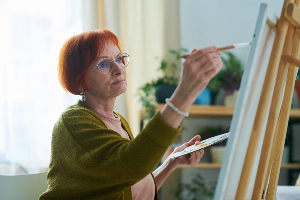 Active senior woman holding palette and paintbrush and painting on canvas in room at home.