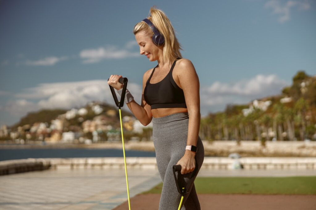 Fitness blonde female listen to music through headphones and strengthen hand muscles while exercising with resistance strap outdoors