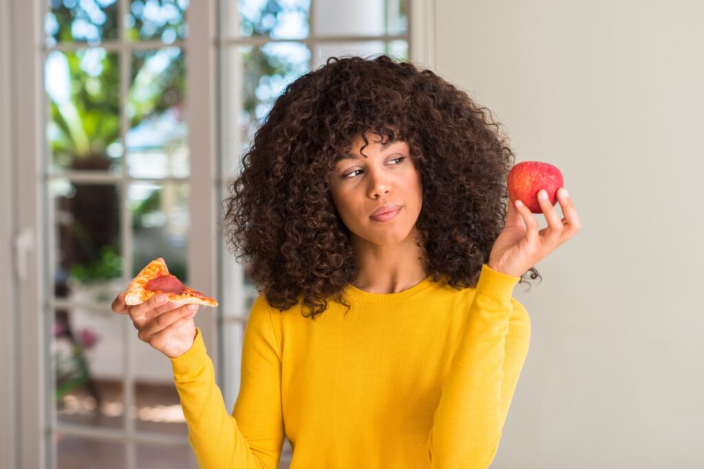 African American woman choosing between apple and pizza slice.