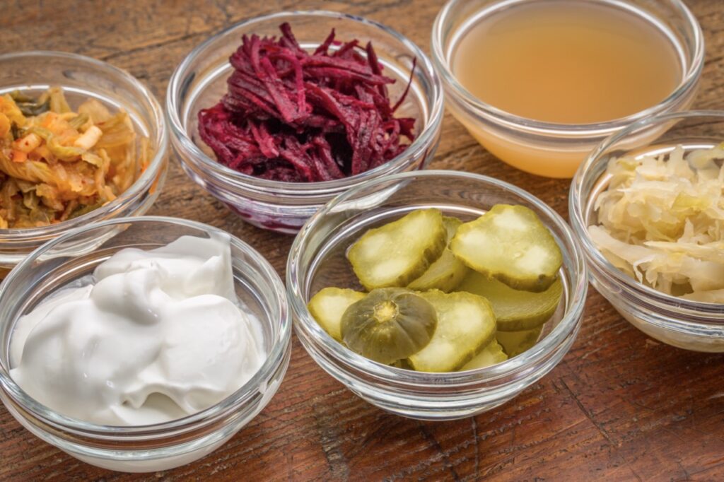 A sampler of fermented, prebiotics, food great for gut health - glass bowls against wood:  kimchi, red beets, apple cider vinegar, coconut milk yogurt, cucumber pickles, sauerkraut.