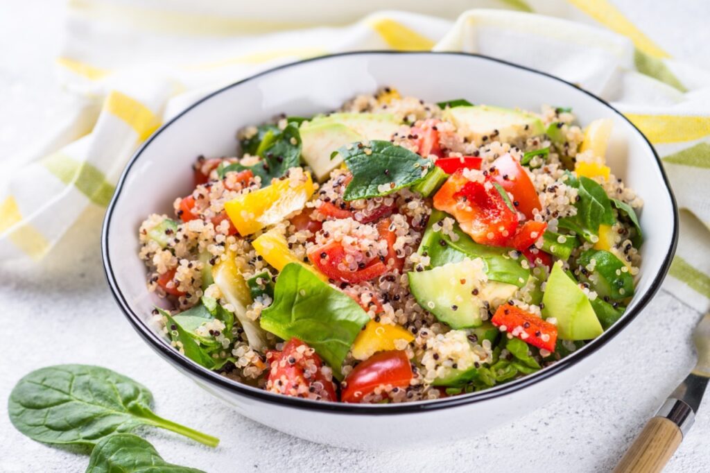 Quinoa salad with spinach, avocado, paprika and tomatoes on white stone table.