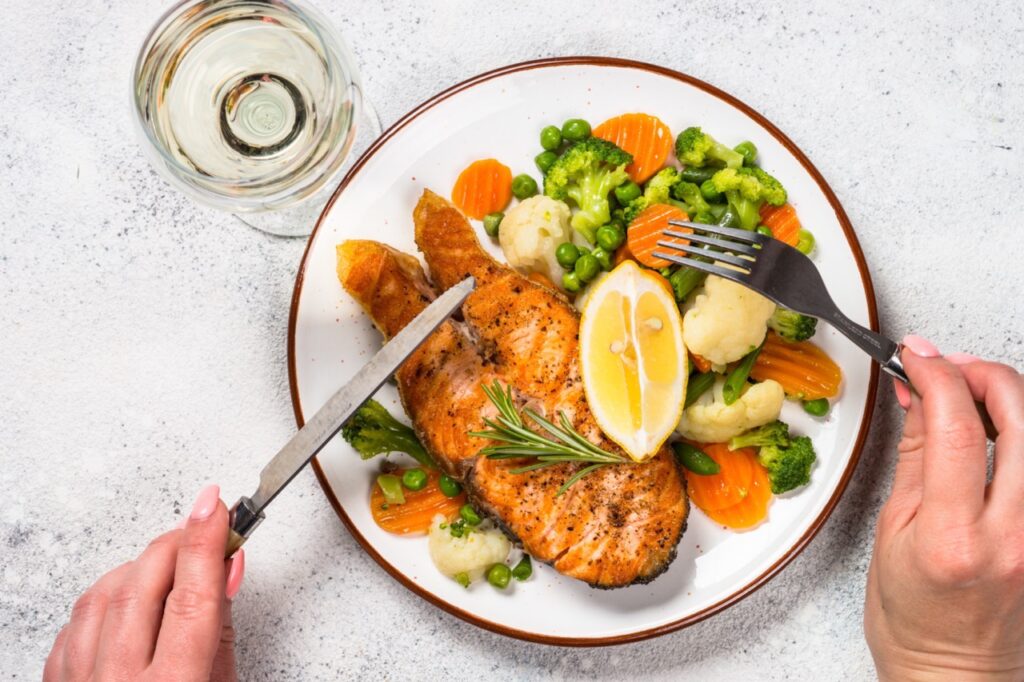 Keto lunch dish. Woman eating Grilled salmon  steak with vegetables  on white stone table. 