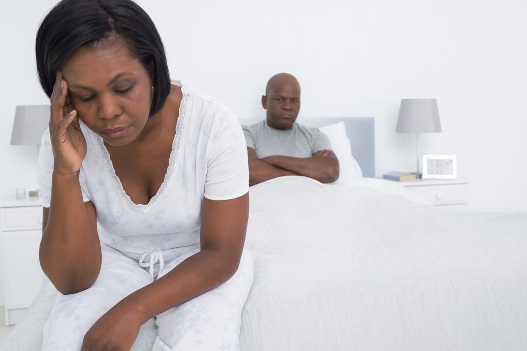 A woman and man sitting on a bed dealing with the frustration of the challenges of menopause.