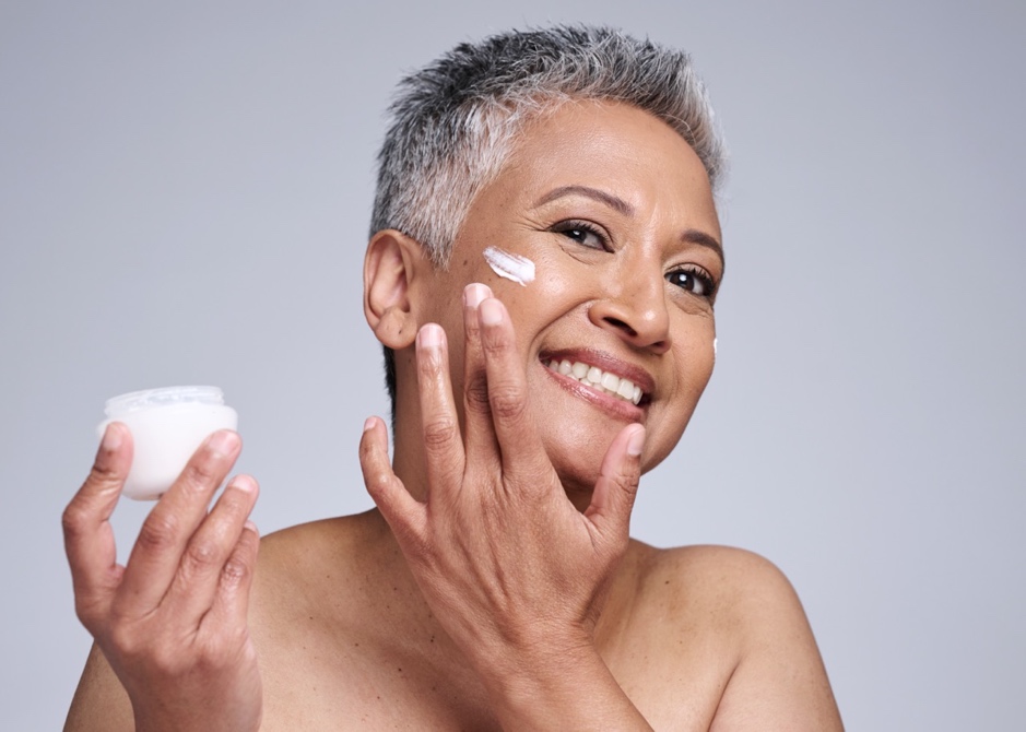Smiling mature woman applying moisturizer to her face, highlighting a hydrating skincare routine for healthy menopausal skin