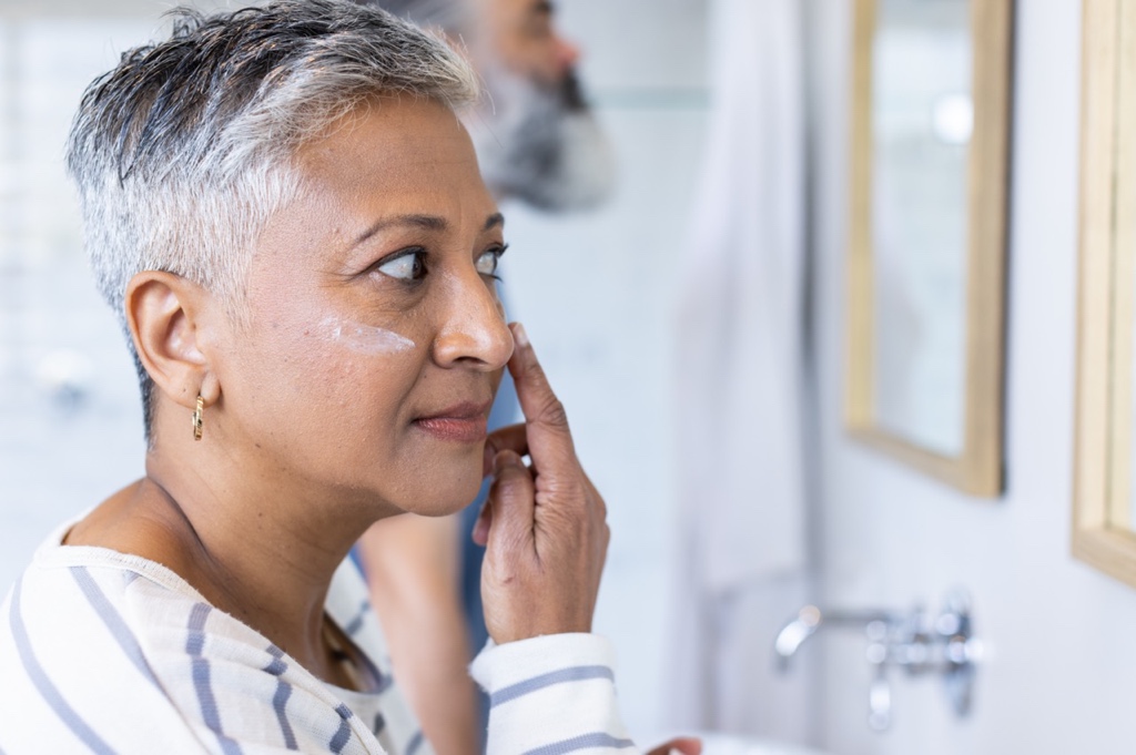 Mature woman applying face cream while looking in a mirror, demonstrating a daily skincare routine to support healthy skin during menopause